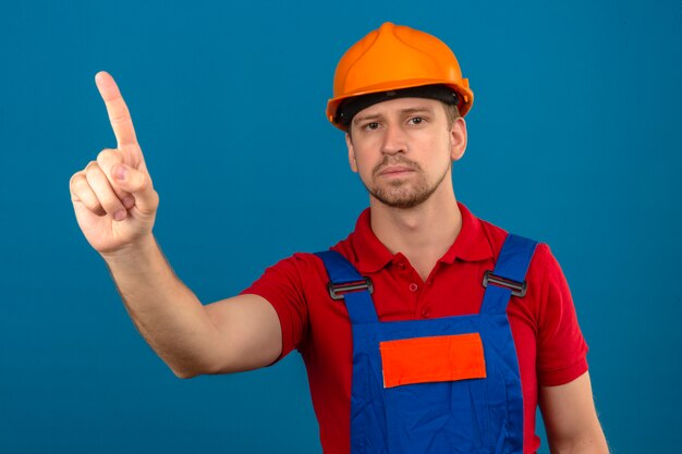 Young builder man in construction uniform and safety helmet pointing with finger up with serious face over isolated blue wall