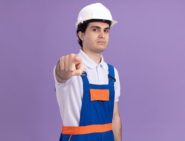 Young builder man in construction uniform and safety helmet pointing with index finger at front looking displeased standing over purple wall