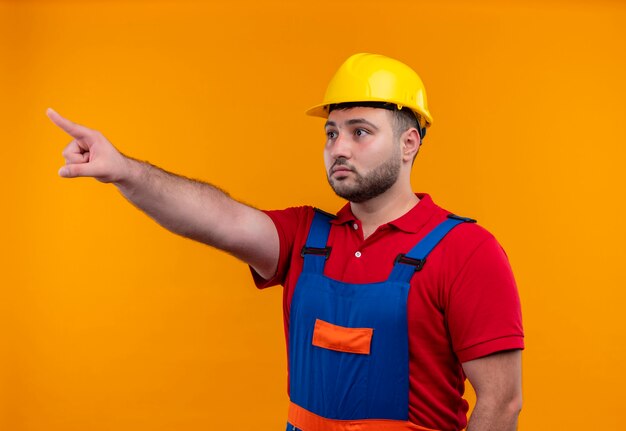 Young builder man in construction uniform and safety helmet pointing with index finger to something looking surprised 