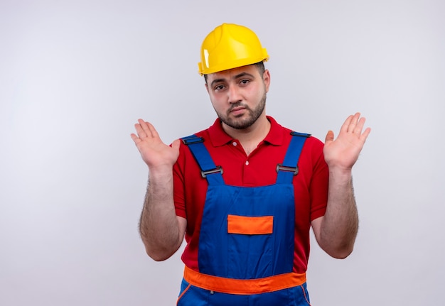 Young builder man in construction uniform and safety helmet raising hands in surrender with sad expression 