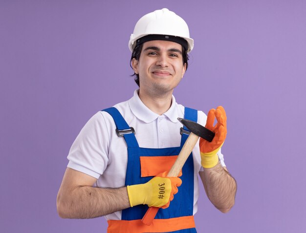 Young builder man in construction uniform and safety helmet in rubber gloves holding hammer looking at front with smile on face standing over purple wall