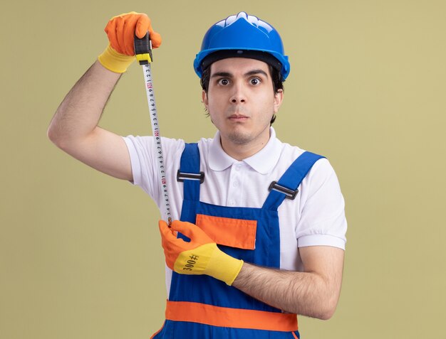 Young builder man in construction uniform and safety helmet in rubber gloves holding measure tape looking at front confused standing over green wall