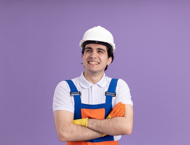 young builder man in construction uniform and safety helmet in rubber gloves looking aside smiling cheerfully with arms crossed standing over purple wall