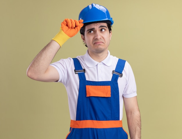 Young builder man in construction uniform and safety helmet in rubber gloves looking confused touching his helmet standing over green wall