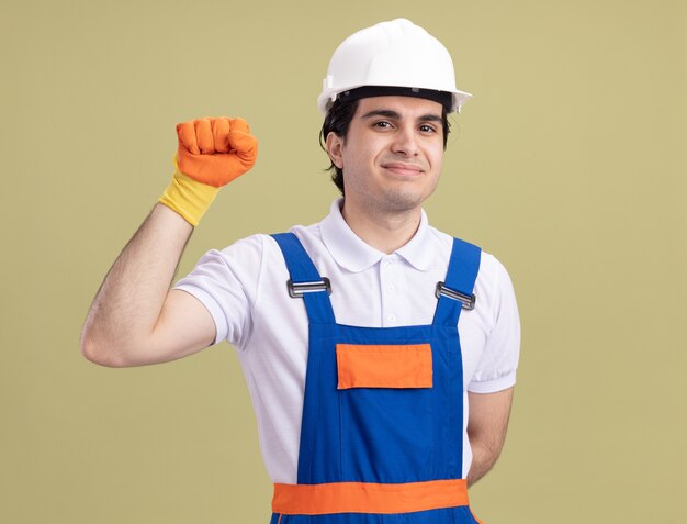 Young builder man in construction uniform and safety helmet in rubber gloves looking at front smiling confident raising fist standing over green wall