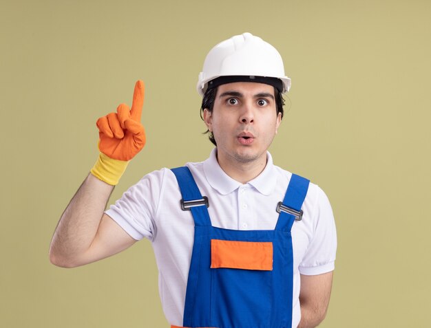 Молодой строитель в строительной форме и защитном шлеме в резиновых перчатках, глядя вперед, удивился, показывая указательный палец, имеющий новую идею, стоящий над зеленой стеной