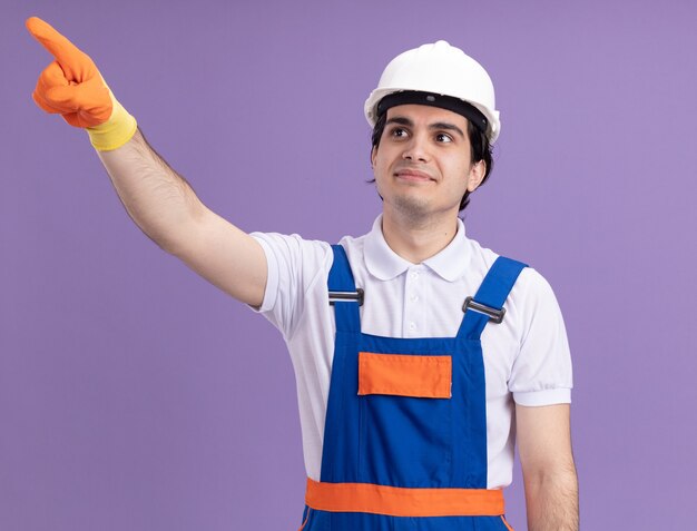 紫色の壁の上に立って微笑んでいる何かに人差し指で指しているゴム手袋の建設制服と安全ヘルメットの若いビルダー男