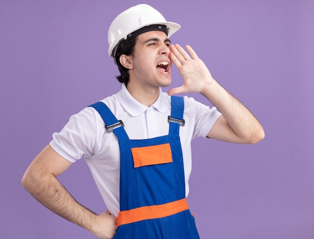 Young builder man in construction uniform and safety helmet shouting or calling someone with hand near his mouth standing over purple wall