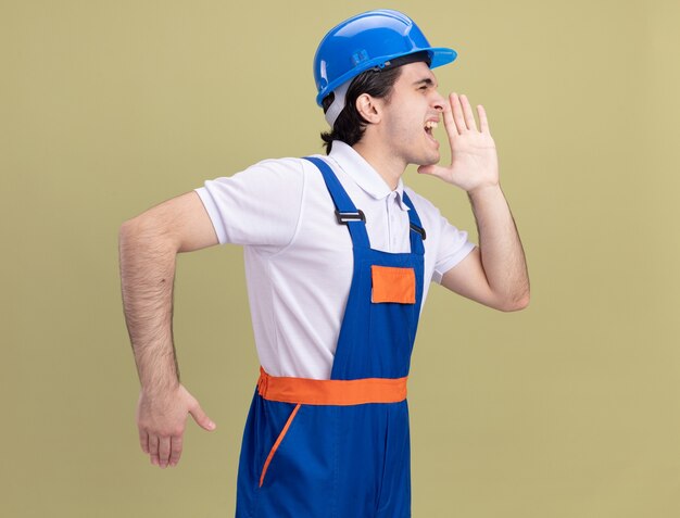 건설 유니폼 및 안전 헬멧에 젊은 작성기 남자 소리 또는 녹색 벽 위에 서있는 입 근처 손으로 누군가를 호출