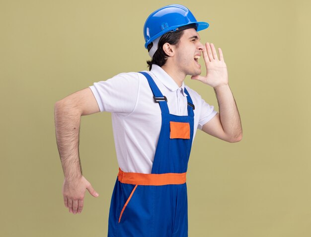 Young builder man in construction uniform and safety helmet shouting or calling someone with hand near mouth standing over green wall