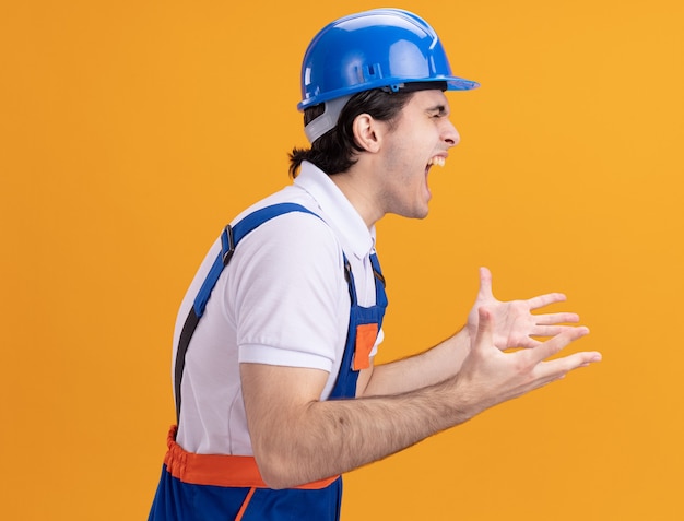 Uomo giovane costruttore in uniforme da costruzione e casco di sicurezza che grida con espressione aggressiva in piedi sopra la parete arancione