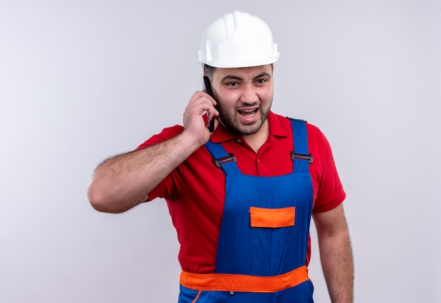 Young builder man in construction uniform and safety helmet shouting with aggressive expression while talking on mobile phone 