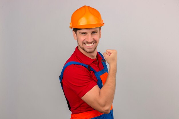 Young builder man in construction uniform and safety helmet showing biceps on his hand smiling winner concept over isolated white wall