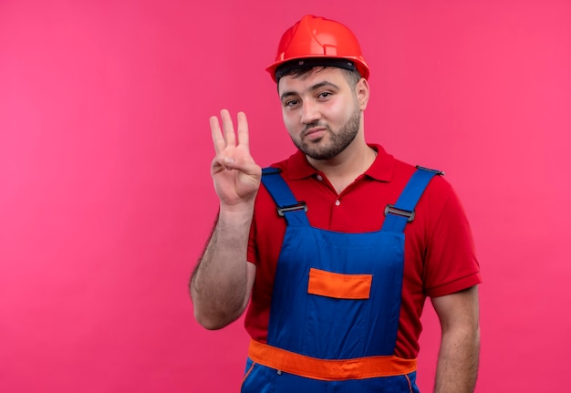 Молодой строитель в строительной форме и защитном шлеме показывает пальцами номер три и показывает вверх