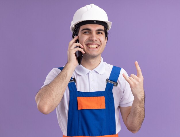 Uomo giovane costruttore in uniforme da costruzione e casco di sicurezza che sorride allegramente facendo simbolo di roccia parlando al telefono cellulare in piedi sopra la parete viola