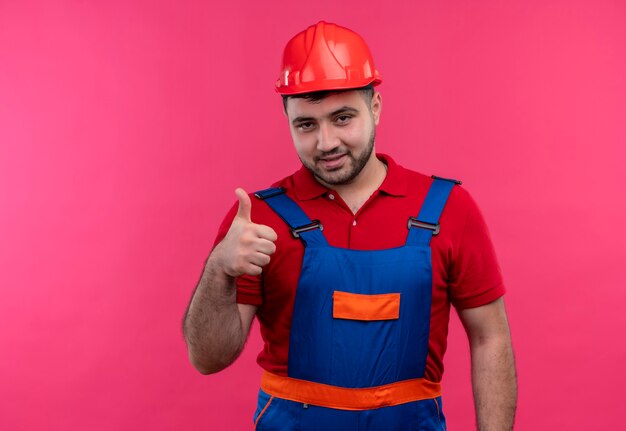 Young builder man in construction uniform and safety helmet smiling cheerfully showing thumbs up 