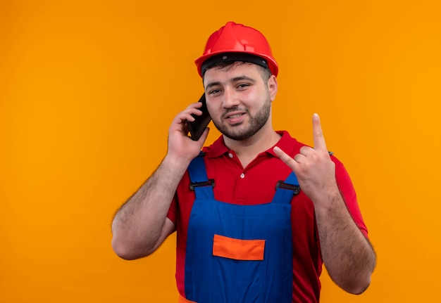 건설 유니폼 및 안전 헬멧에 젊은 작성기 남자는 휴대 전화에 얘기하는 동안 손가락으로 바위 기호 만들기 미소