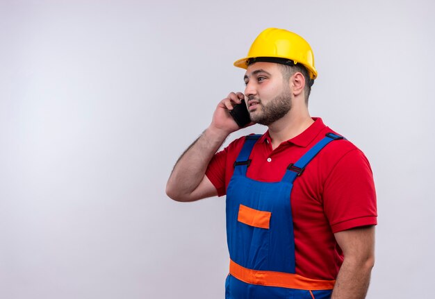 Young builder man in construction uniform and safety helmet smiling while talking on mobile phone 