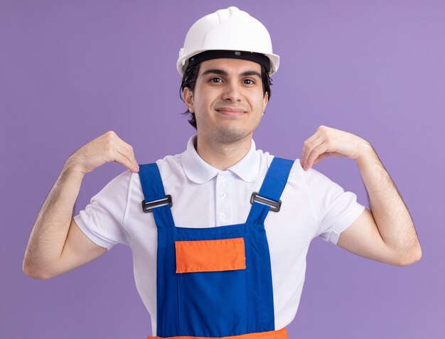 Uomo del giovane costruttore in uniforme da costruzione e casco di sicurezza che sorride con la faccia felice che solleva le mani che tocca le sue spalle che stanno sopra la parete viola