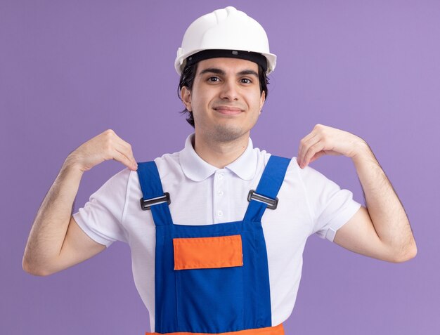 Молодой строитель в строительной форме и защитном шлеме улыбается со счастливым лицом, поднимая руки, касаясь его плеч, стоящих над фиолетовой стеной