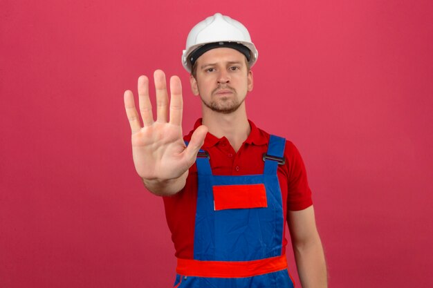 Young builder man in construction uniform and safety helmet standing with open hand doing stop sign with serious and confident expression defense gesture over isolated pink wall