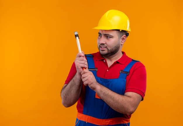 Young builder man in construction uniform and safety helmet swinging a hammer with angry face 