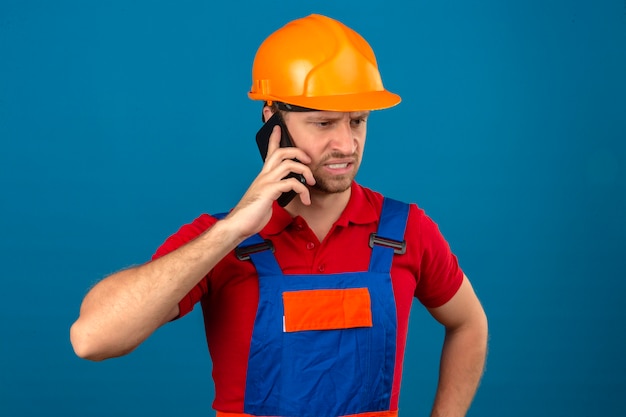 Young builder man in construction uniform and safety helmet talking on mobile phone unhappy in stress over blue isolated wall