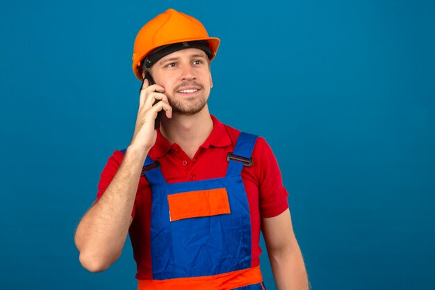 Young builder man in construction uniform and safety helmet talking on mobile phone with happy face smiling over blue isolated wall