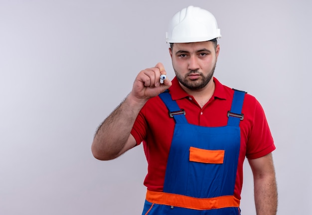 Young builder man in construction uniform and safety helmet trying to write in air with pen looking with serious face 