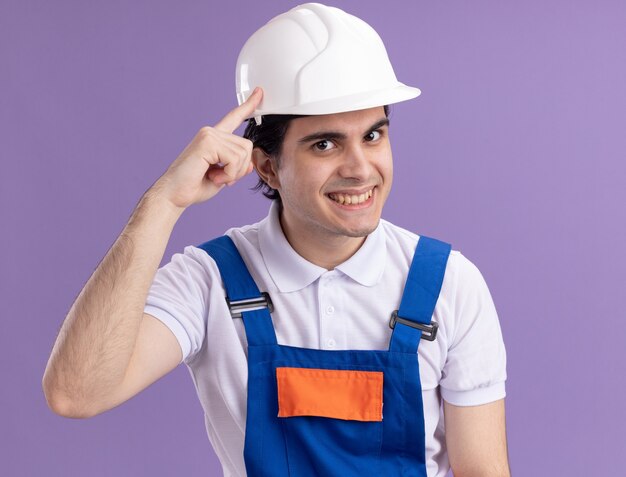 Young builder man in construction uniform and safety helmet  with confident expression on smart face smiling pointing with index finger at his temple focused on a task