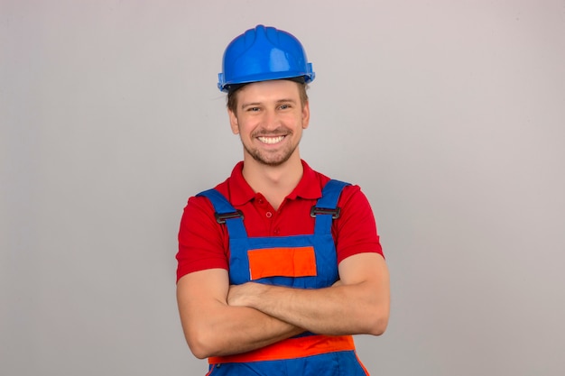 Young builder man in construction uniform and safety helmet with confident smile on face and crossed arms over isolated white wall