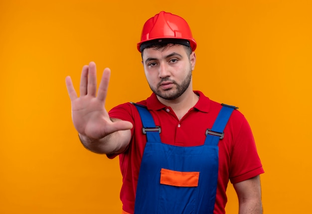 Young builder man in construction uniform and safety helmet  with open hand making stop sign with serious face 