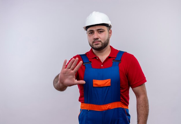Young builder man in construction uniform and safety helmet  with open hand making stop sign with serious face 