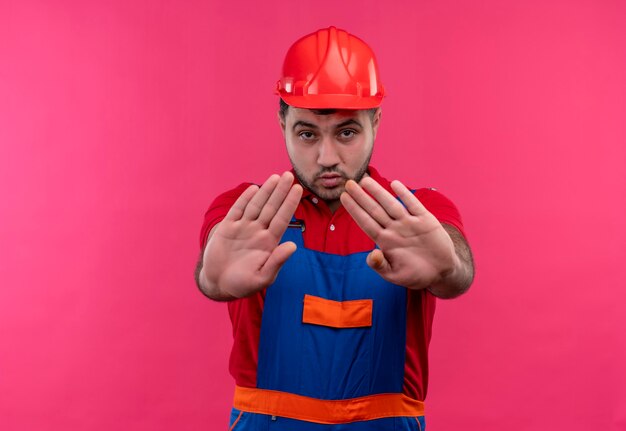 Uomo giovane costruttore in uniforme da costruzione e casco di sicurezza con le mani aperte che fa il fanale di arresto con la faccia seria