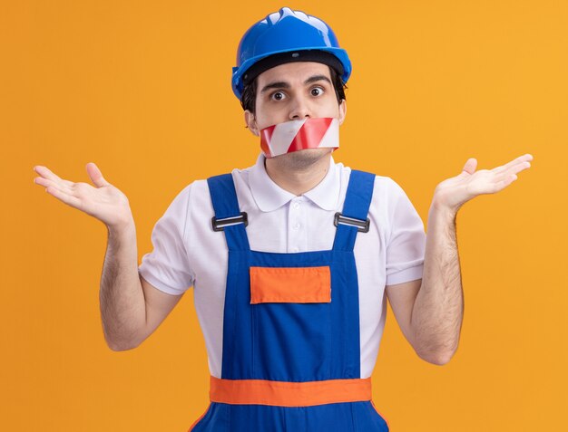 オレンジ色の壁の上に立っている混乱した肩をすくめる肩を見て口の周りにテープを巻いた建設制服と安全ヘルメットの若いビルダー男