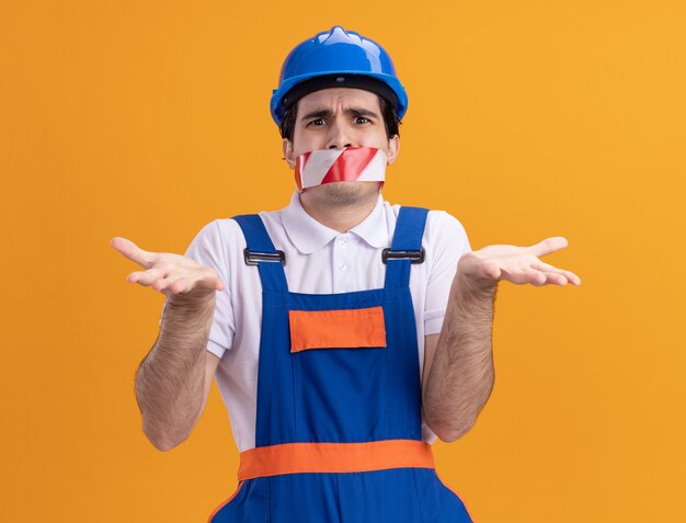 Young builder man in construction uniform and safety helmet with tape wrapped around mouth looking confused shrugging shoulders standing over orange wall