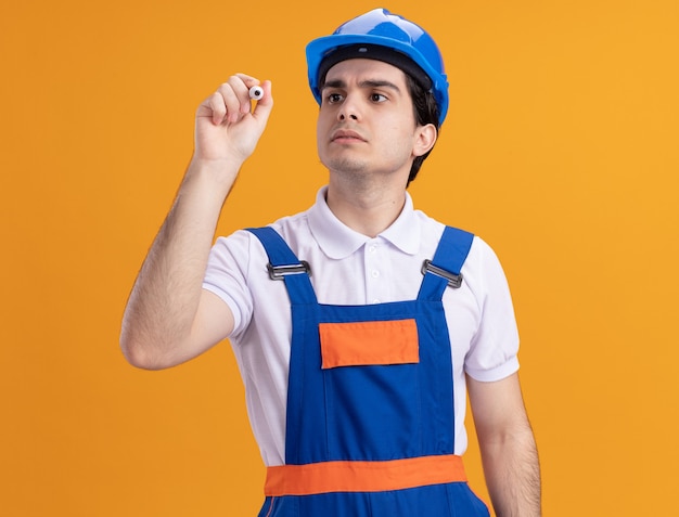 Young builder man in construction uniform and safety helmet writing with pen in front of front with serious face standing over orange wall
