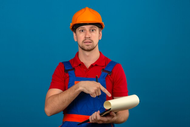 Young builder man in construction uniform and safety helmetyoung builder man in construction uniform and safety helmet with surprised face pointing to clipboard in hands ove