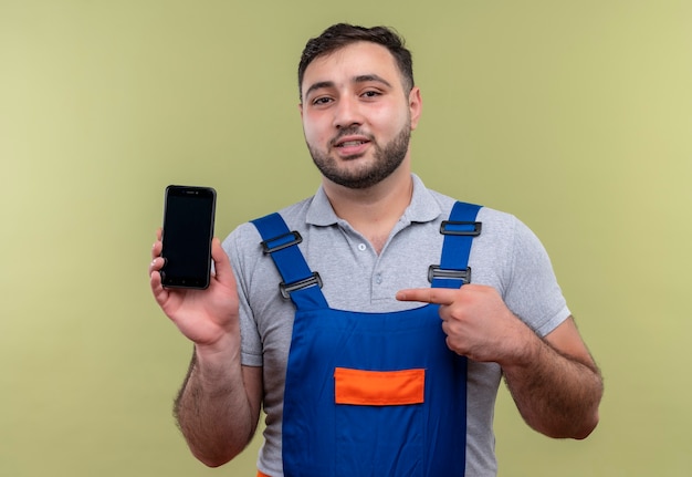 Uomo giovane costruttore in uniforme di costruzione che mostra smartphone che punta con il dito ad esso guardando fiducioso