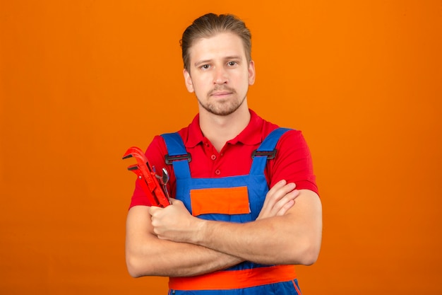 Giovane uomo del costruttore in uniforme della costruzione con il sorriso sicuro sul fronte e le braccia attraversate con la chiave inglese a disposizione sopra la parete arancione isolata