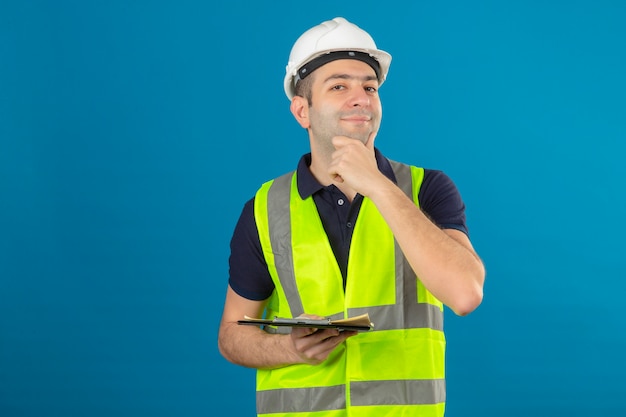Молодой строитель мужчина на мечтательном лице носить белый шлем и желтый жилет с буфером обмена, изолированных на синем