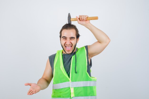 Young builder man holding hammer over head while sticking out tongue in workwear uniform and looking funny. front view.