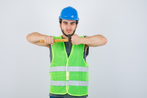 Foto gratuita uomo del giovane costruttore che tiene il martello mentre mostra il pugno chiuso in uniforme da lavoro e che sembra fiducioso. vista frontale.