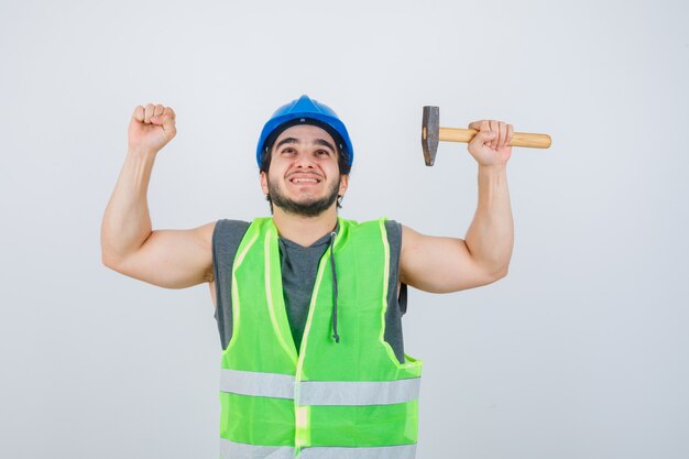 Foto gratuita uomo del giovane costruttore che tiene il martello mentre mostra il gesto del vincitore in uniforme da lavoro e sembra fortunato. vista frontale.