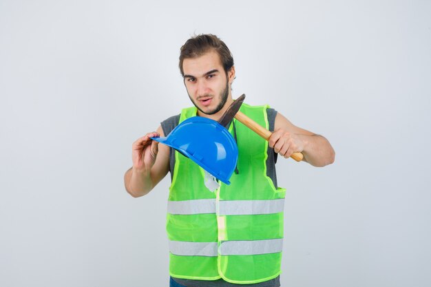 Young builder man holding helmet and hammer in workwear uniform and looking confident. front view.