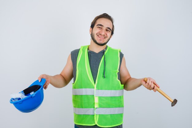 Young builder man holding helmet and hammer in workwear uniform and looking merry , front view.