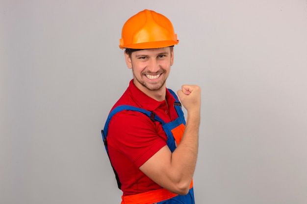 Молодой строитель человек в строительной форме и защитный шлем, показывая бицепс на руке, улыбаясь концепции победителя над изолированной белой стене