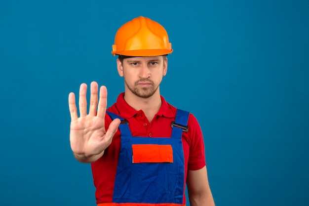 Бесплатное фото Молодой строитель человек в строительной форме и защитный шлем, стоя с открытой руки, делая знак остановки с серьезным и уверенным выражением защиты жест над изолированной синей стеной