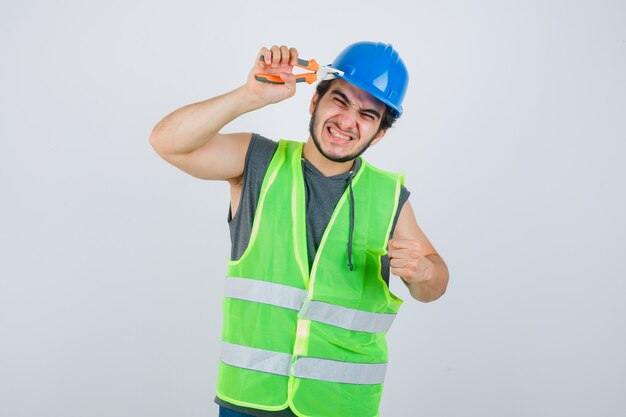 Young builder man pinching helmet with pliers while showing clenched fist in workwear uniform and looking amused. front view.