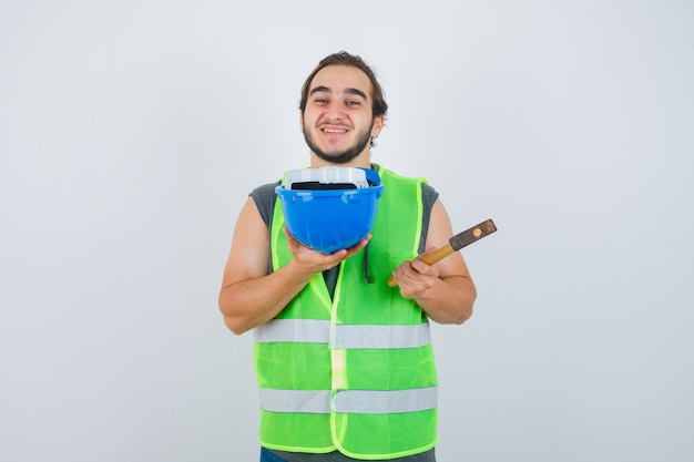 Young builder man showing hammer and helmet in workwear uniform and looking joyful , front view.